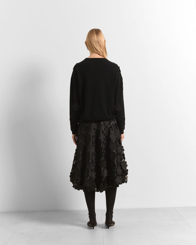 A woman is shown from behind wearing a black long-sleeve knit top and a black textured, ruffled midi skirt with black tights and dark pointed-toe heels, set against a plain white studio background.

Beatrice B- crew neck sweater with embroidered appliques. Available at Edit by Elaine Turner in Houston, TX. Model full back view. 