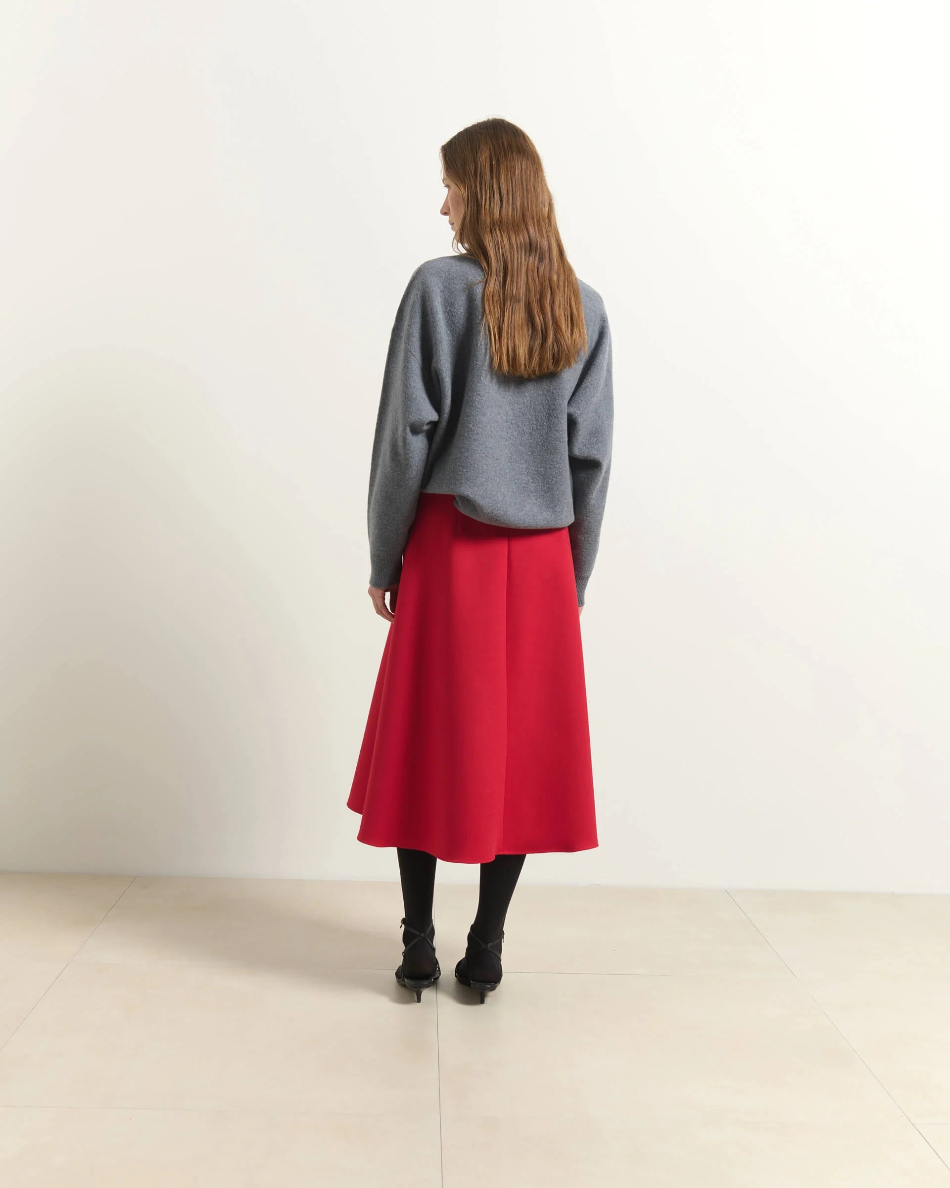A woman stands with her back to the camera wearing a gray knit sweater and a bright red midi skirt, paired with black tights and black ankle boots, in a minimalist studio setting.

Beatrice B Flared Midi Skirt Red. Available at Edit by Elaine Turner in Houston, TX. Full back model view. 
