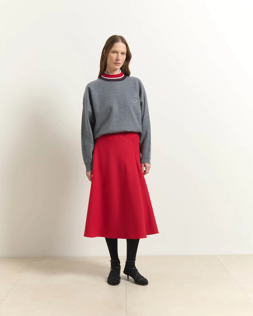 A model stands in a studio wearing a gray knit sweater with a red and navy striped mock neck collar, paired with a bright red A-line midi skirt, black tights, and black heeled boots.

Beatrice B Flared Midi Skirt Red. Available at Edit by Elaine Turner in Houston, TX. Full front model view. 