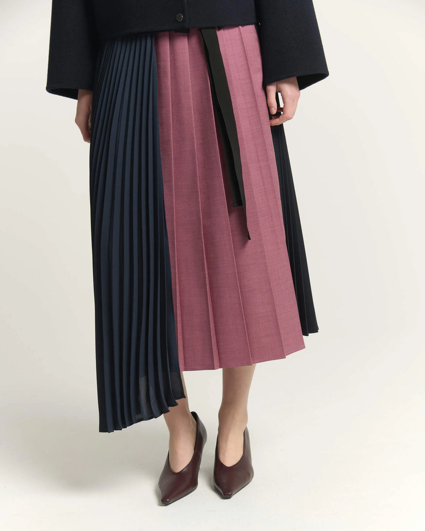 A model is shown from the waist down wearing a pleated midi skirt with three distinct vertical color-blocked panels: navy blue, dusty rose pink, and black. The skirt is styled with a black waist tie and paired with dark brown pointed-toe flats against a clean, off-white studio background.

Beatrice B Pleated Midi Skirt with Panels in Raspberry Pink. Available at Edit by Elaine Turner in Houston, TX. Model close up View. 
