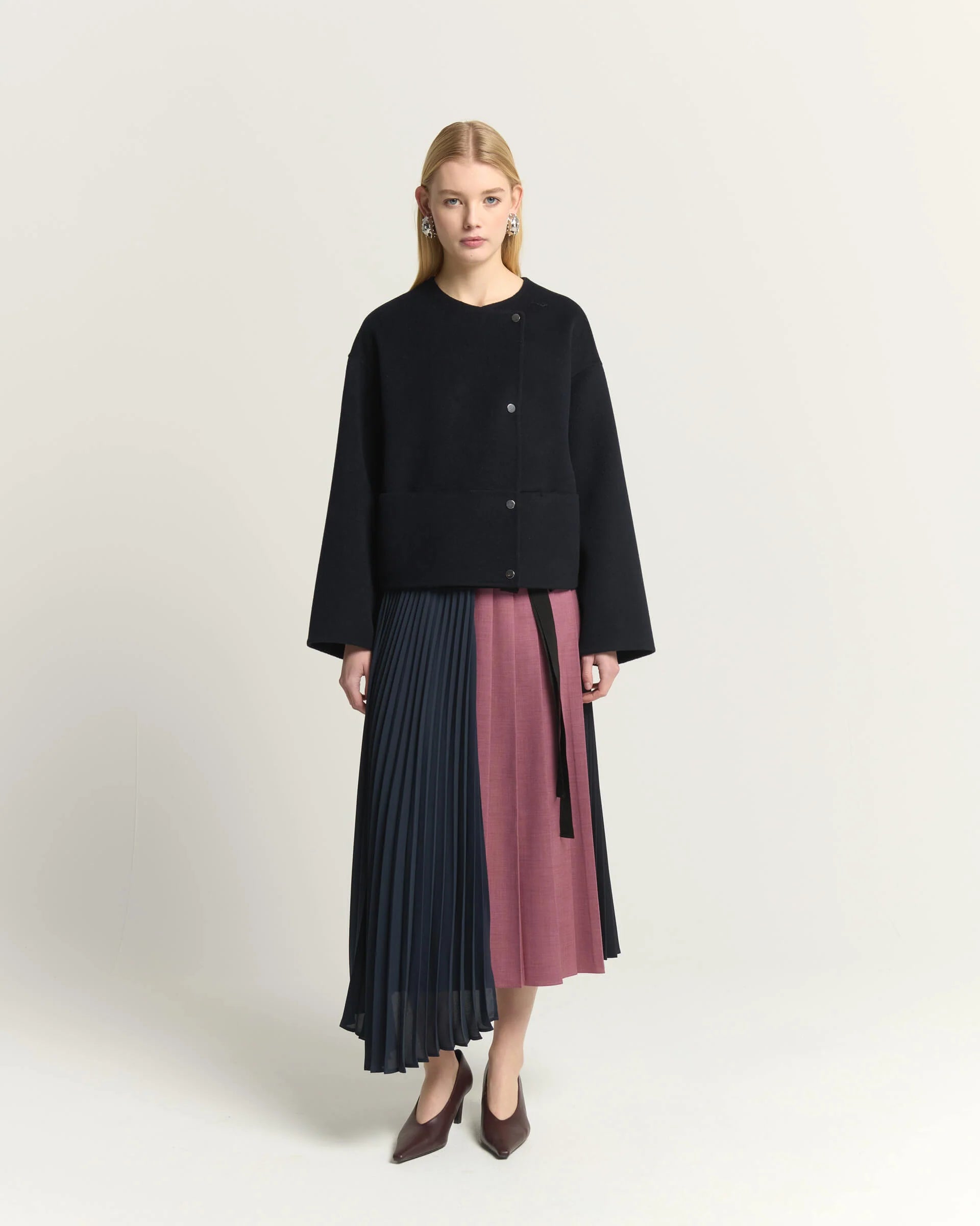 A model stands against a light beige studio background wearing a black cropped cardigan and a pleated midi skirt with color-blocked panels in dark navy and dusty rose. She is wearing dark brown pointed-toe heels.

Beatrice B Pleated Midi Skirt with Panels in Raspberry Pink. Available at Edit by Elaine Turner in Houston, TX. Full front Model View. 