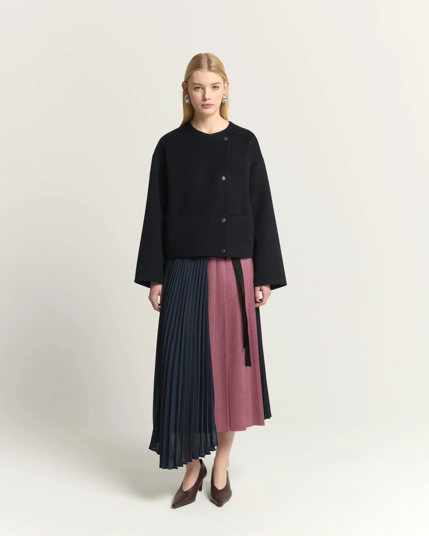 A model stands against a light beige studio background wearing a black cropped cardigan and a pleated midi skirt with color-blocked panels in dark navy and dusty rose. She is wearing dark brown pointed-toe heels.

Beatrice B Pleated Midi Skirt with Panels in Raspberry Pink. Available at Edit by Elaine Turner in Houston, TX. Full front Model View. 