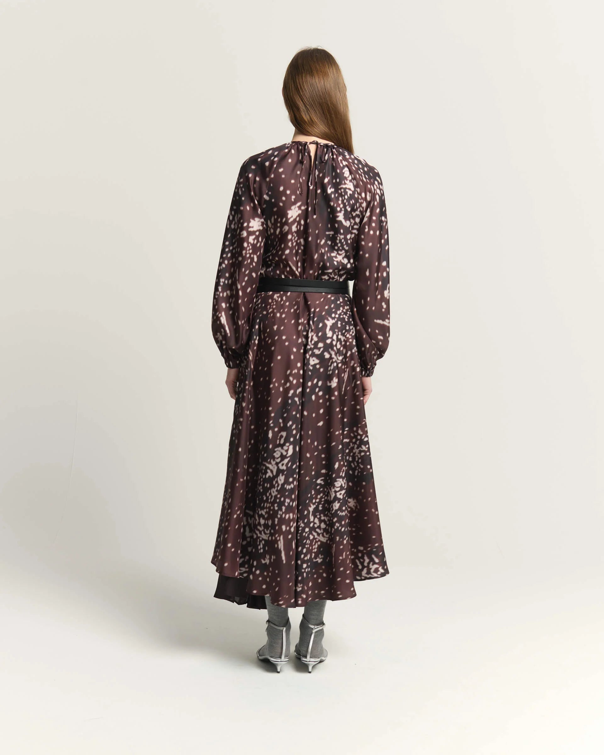 A woman viewed from the back wearing a long-sleeve midi dress with a burnt amber abstract floral pattern, a black waist belt, and gray pointed-toe heels, set against a plain light gray studio background.

Beatrice B Printed Silk Midi Dress with Pleats. Available at Edit by Elaine Turner in Houston, TX. Full back model view. 