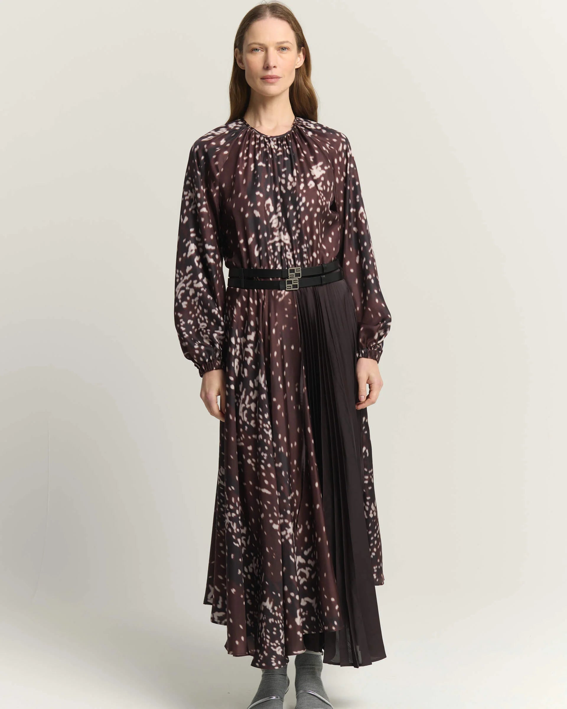 A woman models a long-sleeved midi dress with a burnt amber abstract print, featuring a tie-neck detail, a matching black belt, and a pleated skirt, set against a plain light gray background.

Beatrice B Printed Silk Midi Dress with Pleats. Available at Edit by Elaine Turner in Houston, TX. Full front model view. 