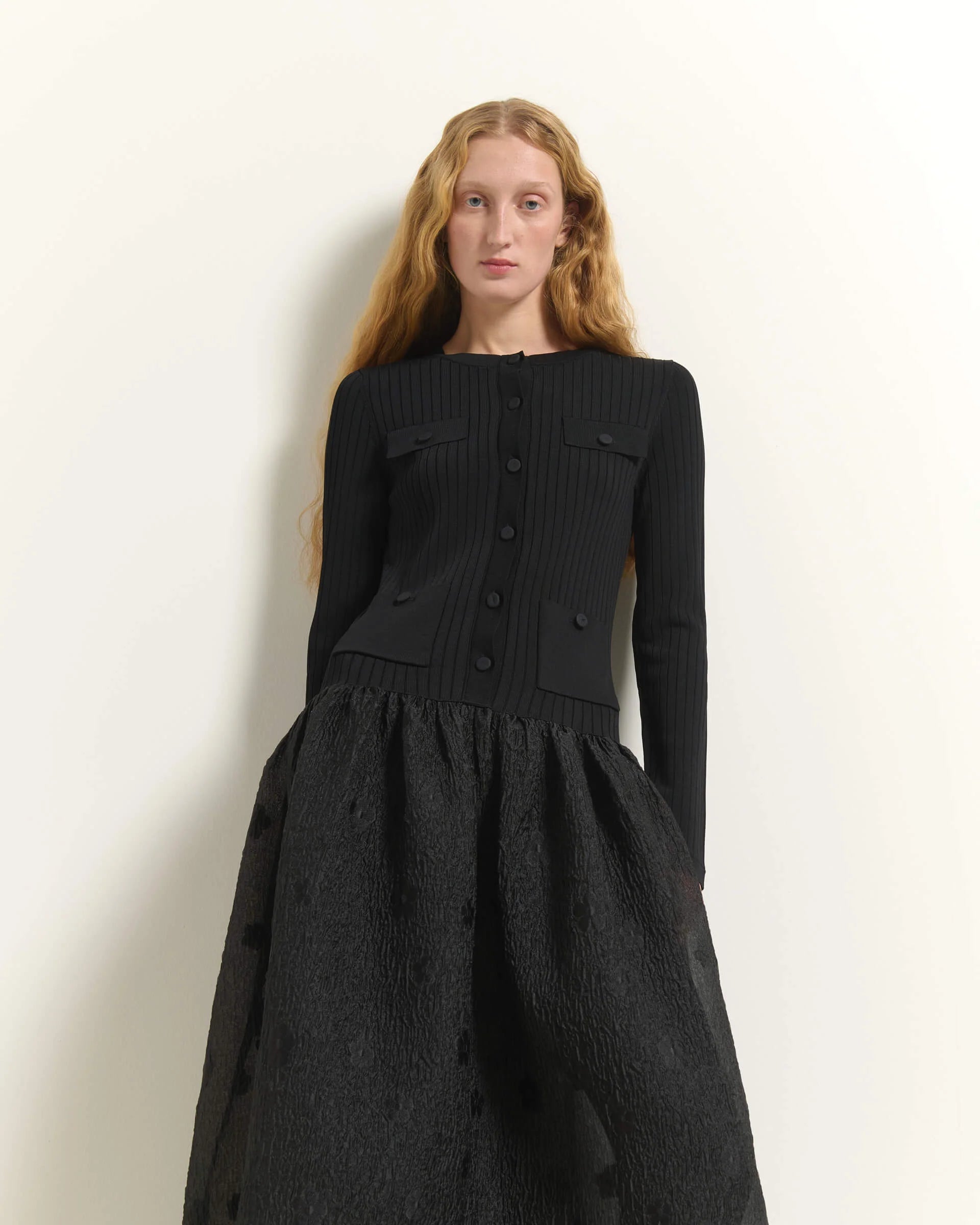 A model with long light-brown hair wearing a black ribbed long-sleeve top with front buttons and a matching textured black skirt, standing against a plain off-white wall.

Beatrice B. Ribbed Knit Dress with Jacquard Skirt in black. Available at Edit by Elaine Turner in Houston, TX. Model close up View.