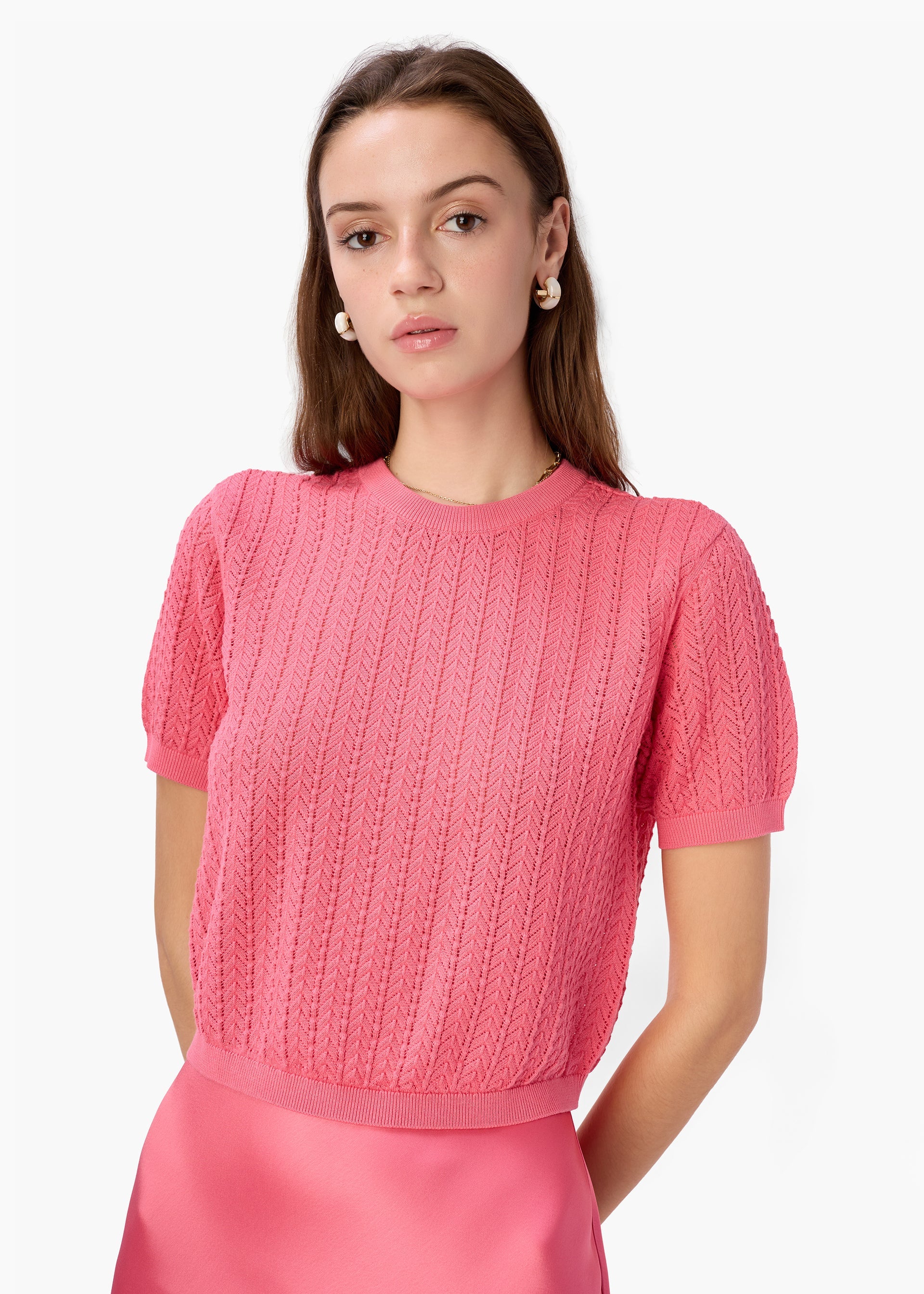 Woman wearing a pink knitted top against a white background by CAMI NYC - Ellory Cotton Knit Top in Hibiscus at edit. BY ELAINE TURNER. Model shown wearing the Ellory from the front view. 