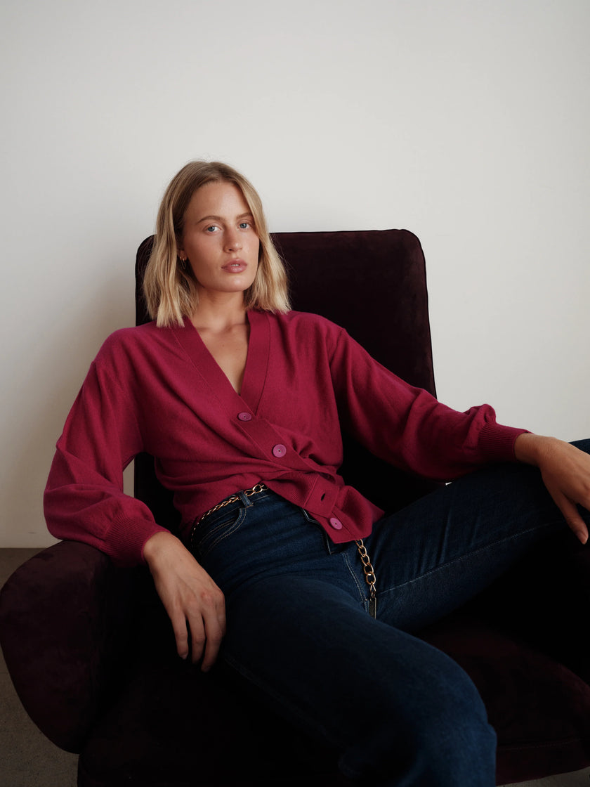 A woman with blonde hair sits in a dark purple armchair, wearing a fuchsia cardigan with a V-neck and front buttons, paired with dark blue jeans.

CISSA- The Slouch Wrap Cardigan in fuchsia. Available at Edit by Elaine Turner in Houston, TX. Front model view. 