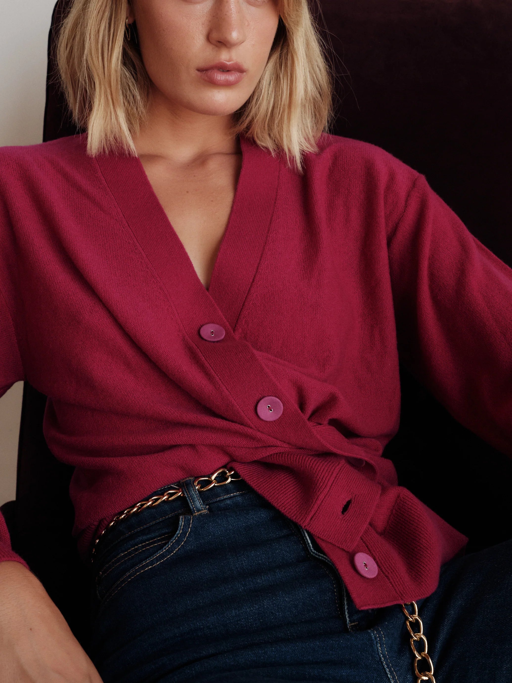 A close-up of a woman wearing a deep fuchsia wrap-style knit cardigan with matching buttons, styled with dark blue jeans and a gold chain belt.

CISSA- The Slouch Wrap Cardigan in fuchsia. Available at Edit by Elaine Turner in Houston, TX. Front model view. 
