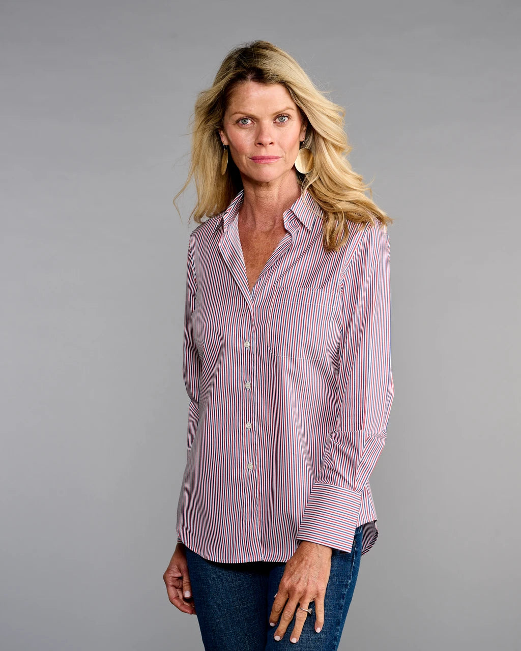 A woman modeling a long-sleeve button-up shirt with thin red and blue vertical stripes on a light pinkish base, paired with dark blue jeans, against a solid gray studio background.

Claridge & King- The 4..0 Shirt in Ross Stripe. Available at Edit by Elaine Turner in Houston, TX. Model Front View 