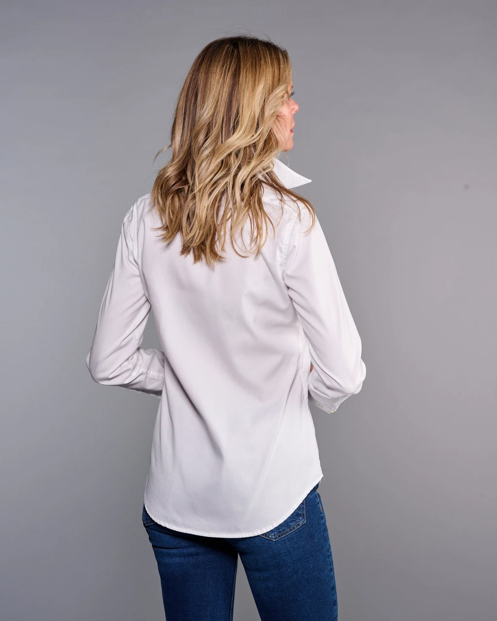 A woman with blonde hair is shown from behind wearing a white long-sleeve button-down shirt and blue jeans, standing against a solid gray studio background.

Claridge & King- The Great White Shirt No. 1 - Available at Edit by Elaine Turner in Houston, TX. Back Model View