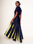 A woman modeling a navy blue short-sleeve maxi dress with bold yellow vertical stripes on the skirt, shown from a three-quarter back angle against a plain off-white studio background.

Daniel Gonzalez Ella Asymmetrical Pleated Dress in Deep Ocean. Available at Edit by Elaine Turner in Houston, TX. Full model back view. 