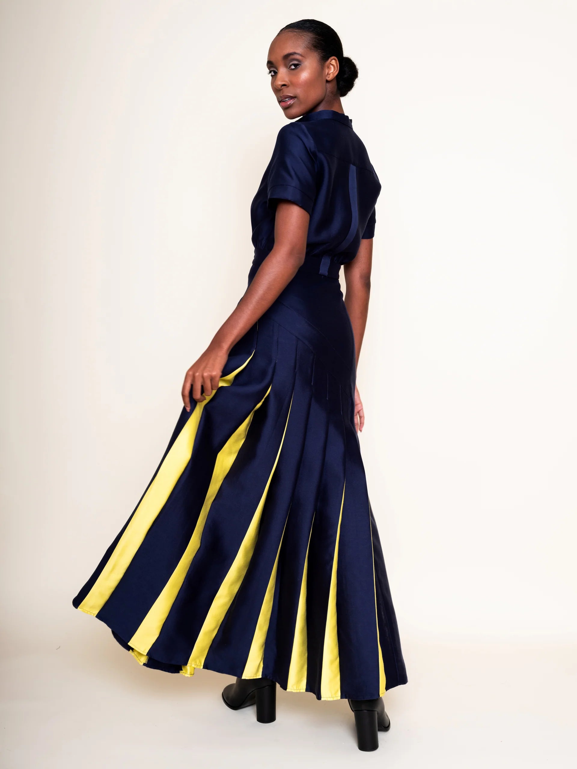 A woman modeling a navy blue short-sleeve maxi dress with bold yellow vertical stripes on the skirt, shown from a three-quarter back angle against a plain off-white studio background.

Daniel Gonzalez Ella Asymmetrical Pleated Dress in Deep Ocean. Available at Edit by Elaine Turner in Houston, TX. Full model back view. 