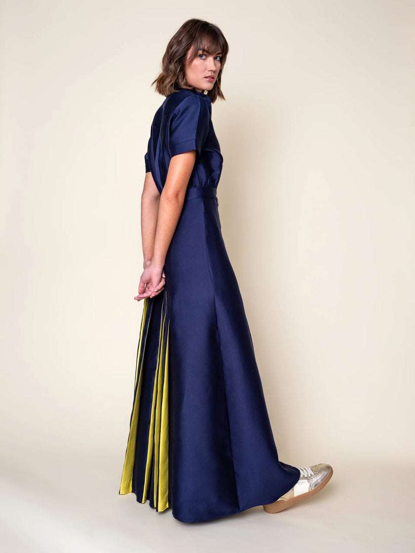 A woman modeling a navy blue silk-cotton blend maxi dress with short sleeves, a high neckline, and a pleated skirt, shown from a three-quarters back view against a solid beige studio background.

Daniel Gonzalez Ella Asymmetrical Pleated Dress in Deep Ocean. Available at Edit by Elaine Turner in Houston, TX. Full model front side view. 