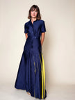 A woman modeling a navy blue silk-cotton blend maxi dress with short sleeves, a buttoned collar, a tie waist, and bold vertical yellow stripes running down the front of the skirt, set against a plain beige studio background.

Daniel Gonzalez Ella Asymmetrical Pleated Dress in Deep Ocean. Available at Edit by Elaine Turner in Houston, TX. Full model front view. 