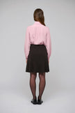 A woman viewed from behind wearing a light pink long-sleeve button-up shirt, a dark brown pleated mini skirt, sheer black tights, and black shoes, standing against a light gray studio background.

Dea Kudibal- Alesandra knit skirt dusty chocolate. Available at Edit by Elaine Turner in Houston, TX.  Model Back View. 