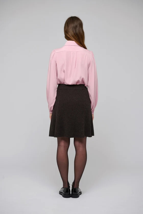 A woman viewed from behind wearing a light pink long-sleeve button-up shirt, a dark brown pleated mini skirt, sheer black tights, and black shoes, standing against a light gray studio background.

Dea Kudibal- Alesandra knit skirt dusty chocolate. Available at Edit by Elaine Turner in Houston, TX.  Model Back View. 