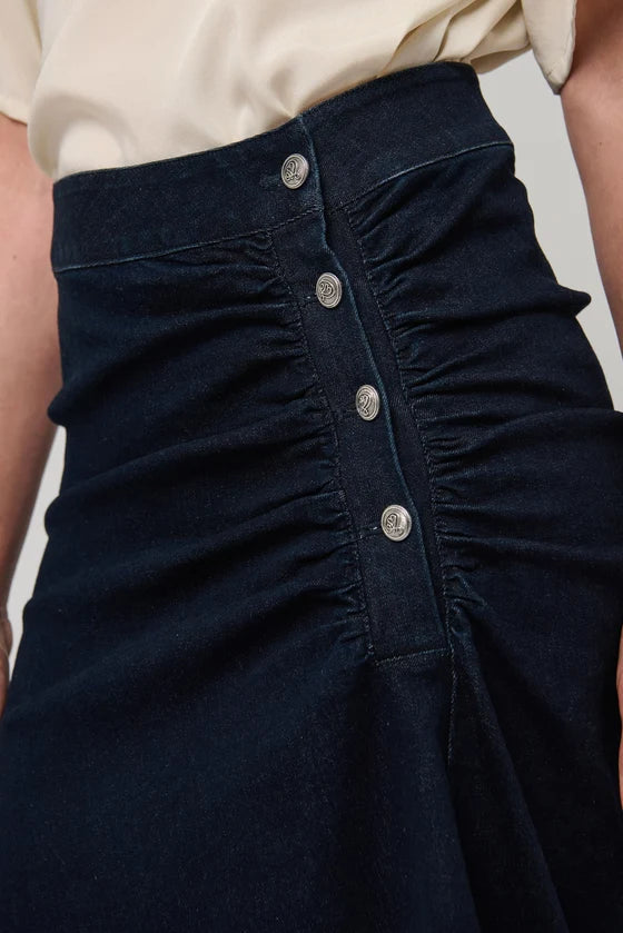Close-up of a dark navy ruched skirt with a button-front closure, worn with a cream top. The fabric features gathered detailing and silver-toned buttons.

Dea Kudibal - Giselle Denim Skirt - Dark Blue. Available at Edit by Elaine Turner in Houston, TX.  Model Close Up View. 