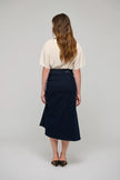 A woman viewed from behind wearing a cream short-sleeve top and a dark navy midi skirt, standing against a light gray studio background.

Dea Kudibal - Giselle Denim Skirt - Dark Blue. Available at Edit by Elaine Turner in Houston, TX.  Model Full Back View. 