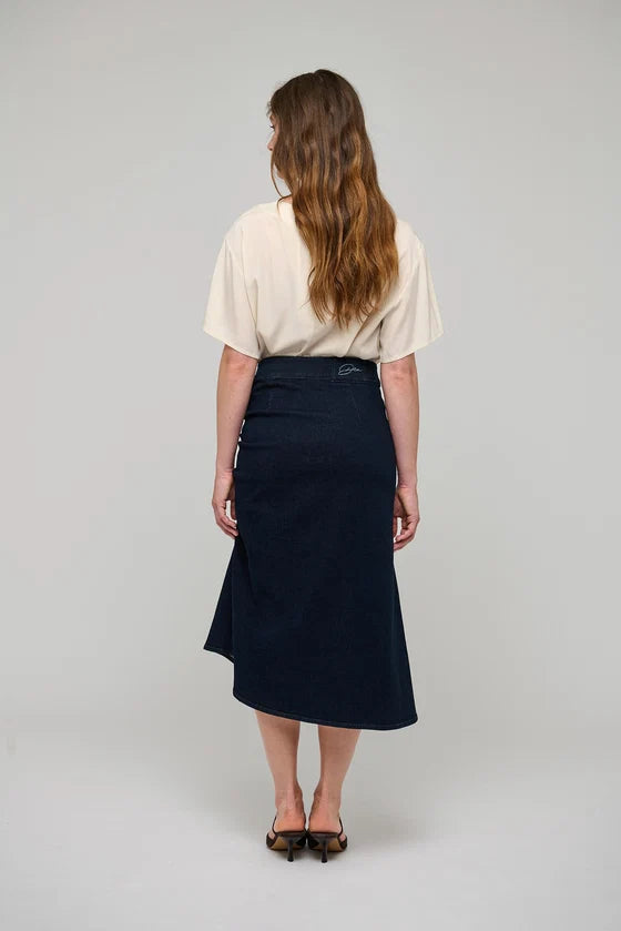 A woman viewed from behind wearing a cream short-sleeve top and a dark navy midi skirt, standing against a light gray studio background.

Dea Kudibal - Giselle Denim Skirt - Dark Blue. Available at Edit by Elaine Turner in Houston, TX.  Model Full Back View. 