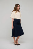 A woman modeling a dark navy midi skirt with a gathered waist and button detail, paired with a cream short-sleeve top and dark brown pointed-toe flats, against a light gray studio background.

Dea Kudibal - Giselle Denim Skirt - Dark Blue. Available at Edit by Elaine Turner in Houston, TX.  Model Full Side View. 