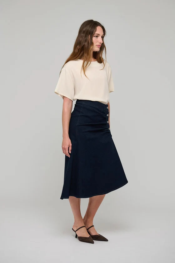 A woman modeling a dark navy midi skirt with a gathered waist and button detail, paired with a cream short-sleeve top and dark brown pointed-toe flats, against a light gray studio background.

Dea Kudibal - Giselle Denim Skirt - Dark Blue. Available at Edit by Elaine Turner in Houston, TX.  Model Full Side View. 