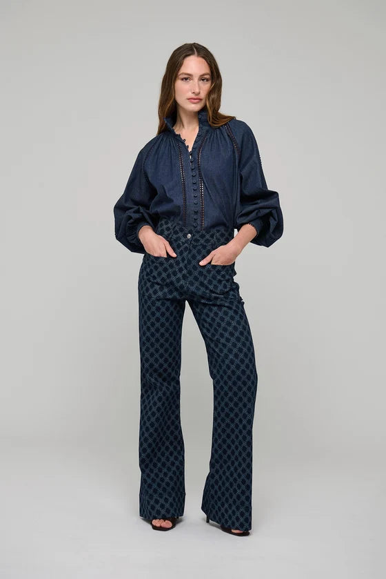 A woman modeling a dark navy blue two-piece set consisting of a long-sleeve button-up blouse with puffed shoulders and matching high-waisted, wide-leg trousers featuring a small geometric pattern, shot against a solid light gray background.

Dea Kudibal Monia Blouse Chambray Blue. Available at Edit by Elaine Turner in Houston, TX . Full Model Front View. 