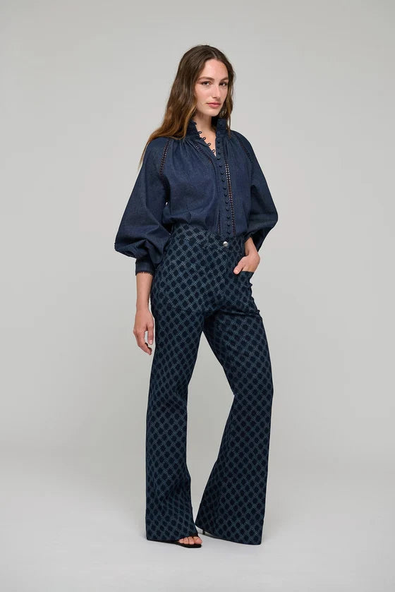 A woman modeling a dark navy blue two-piece outfit consisting of a long-sleeve button-up shirt and matching high-waisted flared trousers with a small geometric pattern, shot against a neutral light gray studio background.

Dea Kudibal Monia Blouse Chambray Blue. Available at Edit by Elaine Turner in Houston, TX . Model Front Side View. 