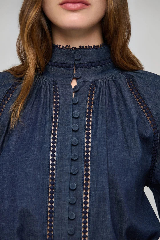Close-up of a model wearing a dark blue denim blouse with a high gathered neckline, front button placket, and decorative geometric embroidered trim.

Dea Kudibal Monia Blouse Chambray Blue. Available at Edit by Elaine Turner in Houston, TX . Model Close Up View. 