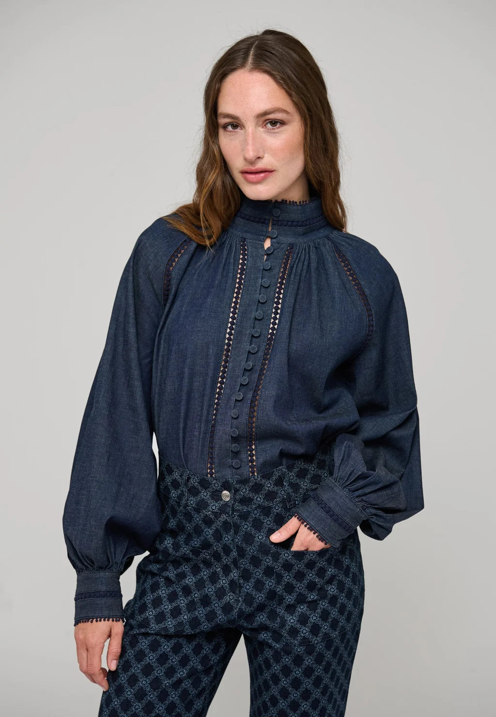 A woman modeling a dark blue long-sleeve blouse with a high gathered neckline, decorative button placket, and contrast stitching, paired with patterned dark pants against a plain light gray studio background.

Dea Kudibal Monia Blouse Chambray Blue. Available at Edit by Elaine Turner in Houston, TX . Model Front View. 
