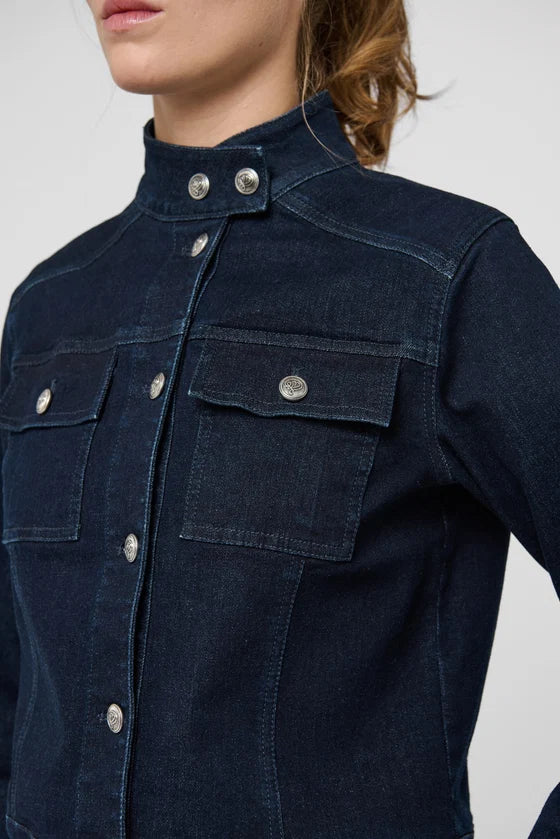 Close-up of a model wearing a dark indigo denim shirt jacket with silver snap buttons and chest pockets, set against a plain light gray background.

Dea Kudibal- Wilma Denim Jacket Dark Blue. Available at Edit by Elaine Turner in Houston, TX. Model Close Up view. 