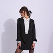 A woman models a black long-sleeve mini dress with a white shirt collar and button placket, trimmed with black fringe at the collar and cuffs, set against a plain light background.

Edward Achour Extended Length Shift Dress with Contrast Bib in Black. Available at Edit by Elaine Turner in Houston, TX. Model front view. 