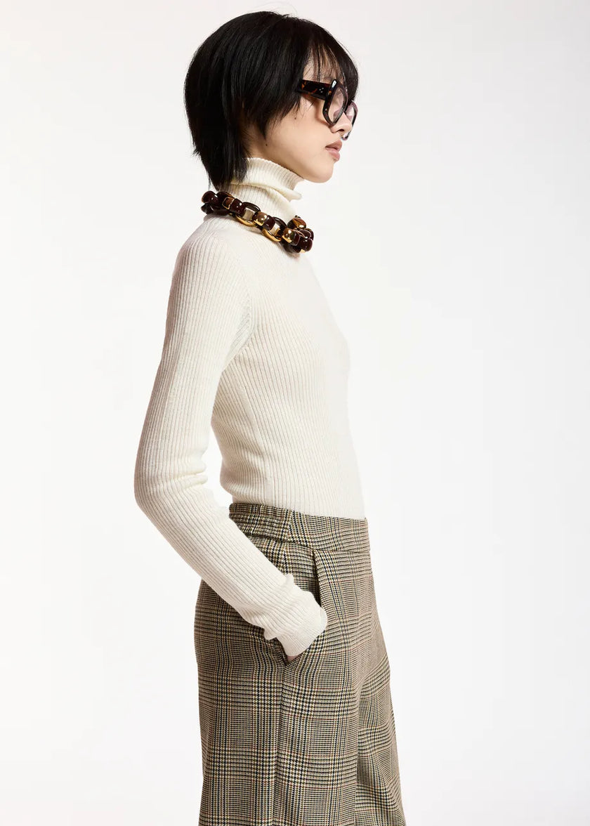 A model in profile wearing a cream ribbed turtleneck sweater, brown plaid trousers, and a chunky brown chain necklace, set against a plain white studio background.

Essentiel Antwerp - Kelmuse Knitted Pullover - Off White. Available at Edit by Elaine Turner in Houston, TX. Model side view. 