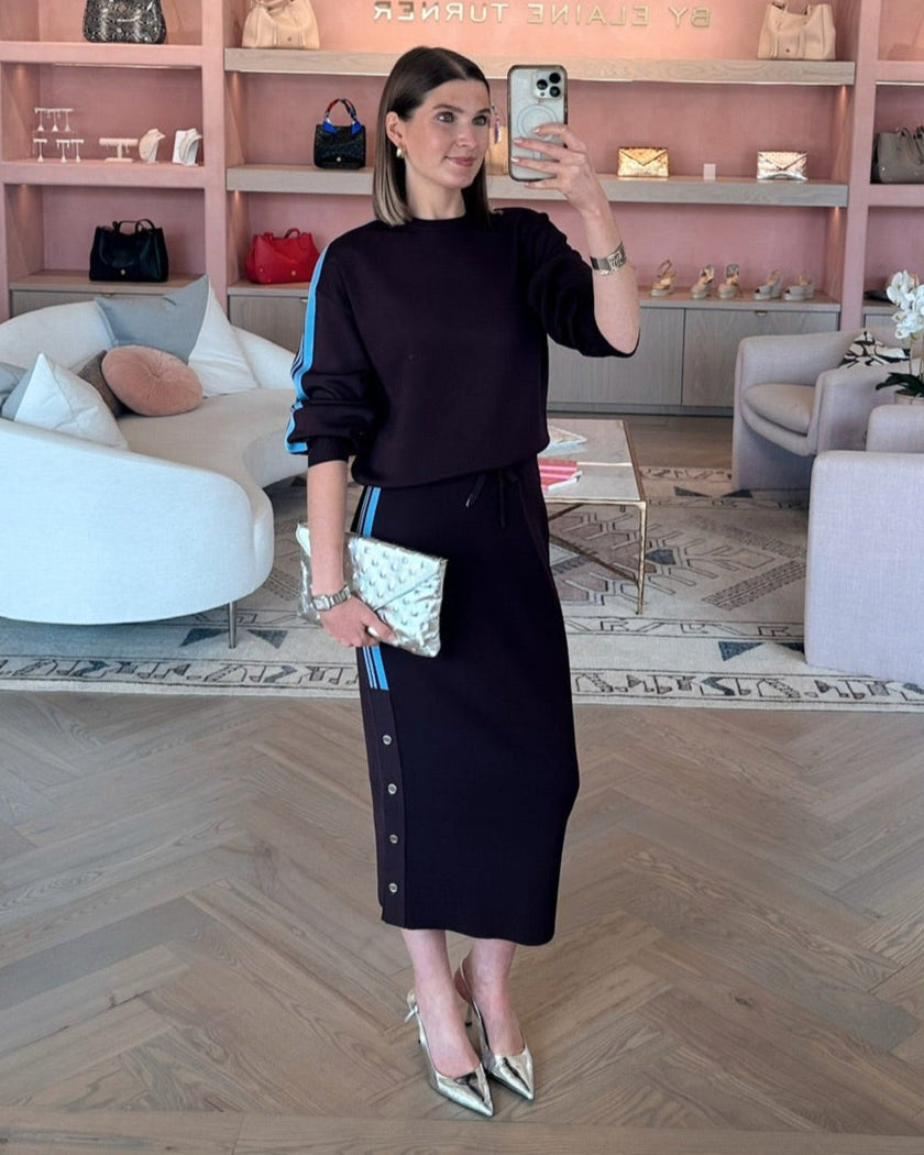 Woman taking a selfie in a stylish interior setting with shelves displaying Elaine Turner handbags wearing a PINKO brand set in Burgandy/ chocolate brown with light blue striped on arm and down side of skirt. 