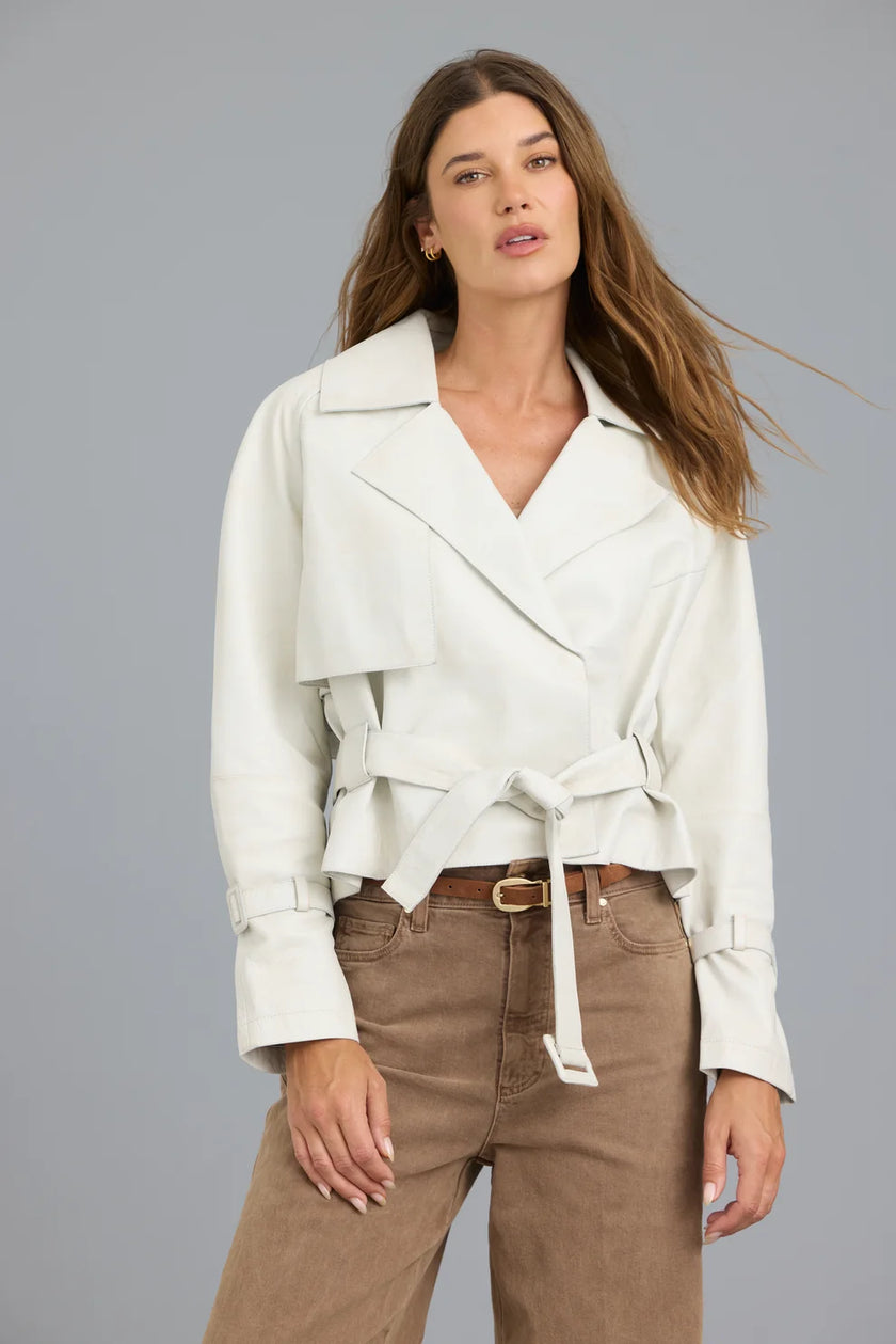 A woman modeling a white wrap-style jacket with a tie belt, paired with brown pants, against a solid gray studio background.

JAKETT- Naomi Vintage Leather Jacket in Coconut. Available at Edit by Elaine Turner in Houston, TX. Front model view. 