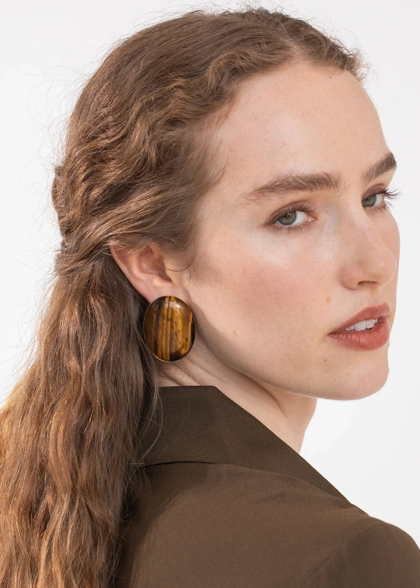 A close-up side profile of a woman with light brown hair wearing a large, round wooden earring with a tiger-stripe pattern, paired with a brown blazer against a plain white background.

Jennifer Behr- Audra Earrings - Tigers Eye. Available at Edit by Elaine Turner in Houston, TX. Model View. 