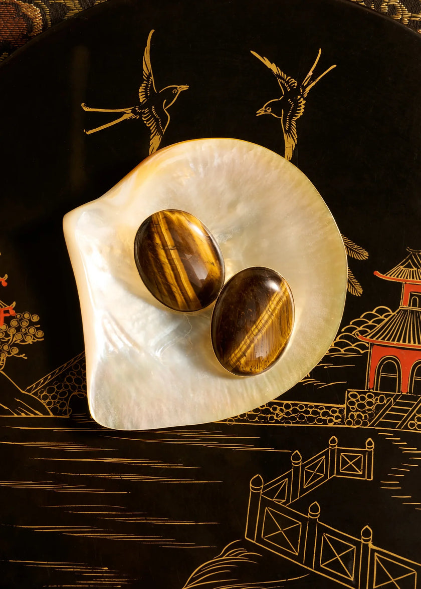 Two polished tiger's eye oval earrings displayed on a white mother-of-pearl shell dish, set against a black decorative background with gold bird and pagoda illustrations.

Jennifer Behr- Audra Earrings - Tigers Eye. Available at Edit by Elaine Turner in Houston, TX. 