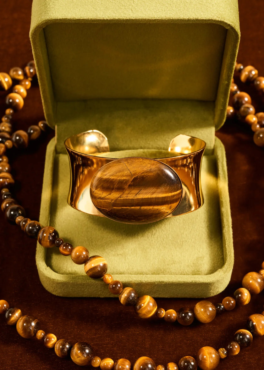 A polished gold cuff bracelet with a large oval tiger's eye stone centerpiece, displayed inside an open olive green velvet jewelry box, surrounded by a matching tiger's eye bead necklace on a dark wooden surface.

Jennifer Behr- Johanna Cuff in Tigers Eye. Available at Edit by Elaine Turner in Houston, TX 