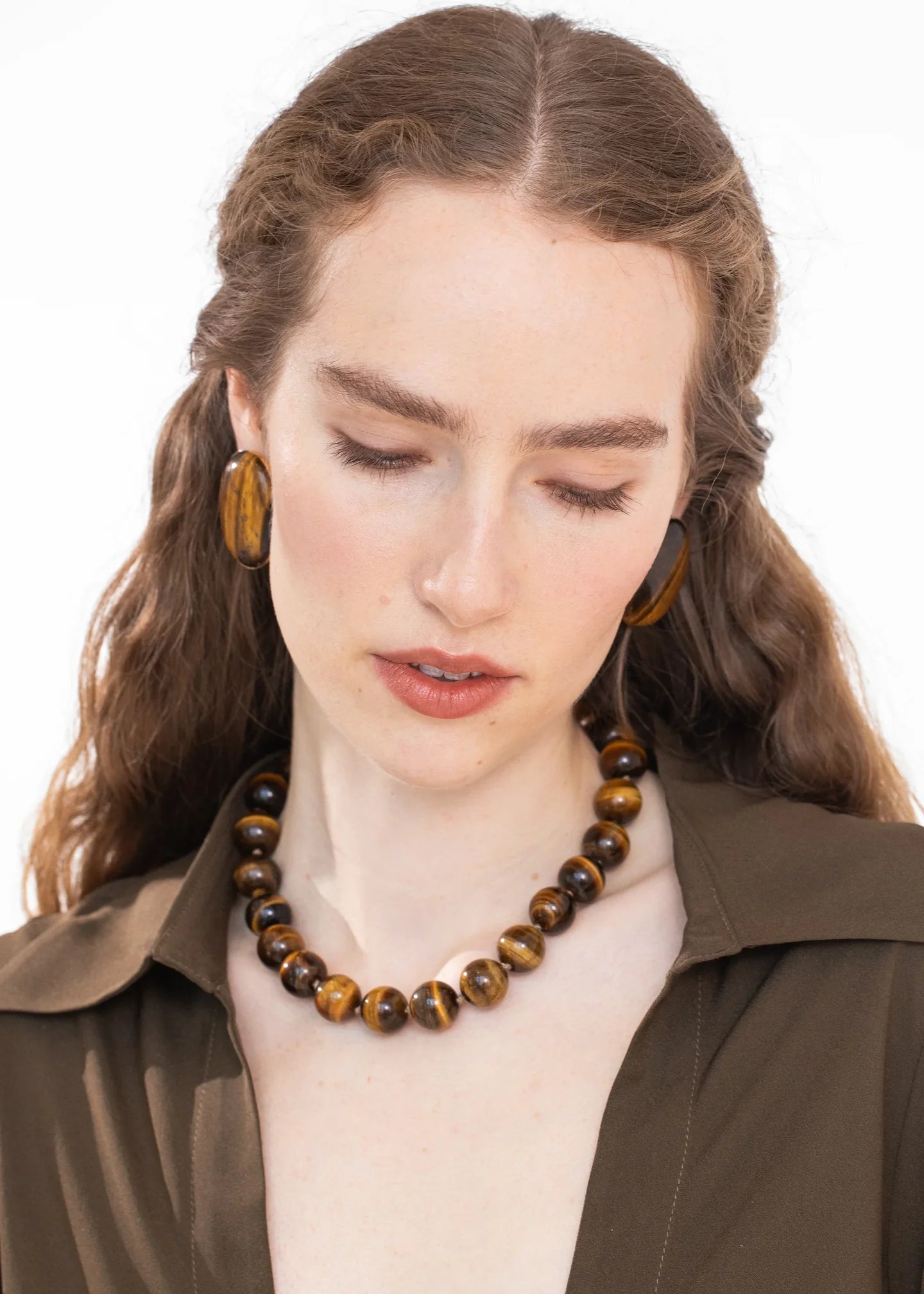 A woman with brown hair wearing a chunky brown and black tiger's eye beaded necklace and matching large round earrings, styled with an olive green shirt against a white background.

Jennifer Behr- Louisa Necklace - Tigers Eye. Available at Edit by Elaine Turner. Model View. 