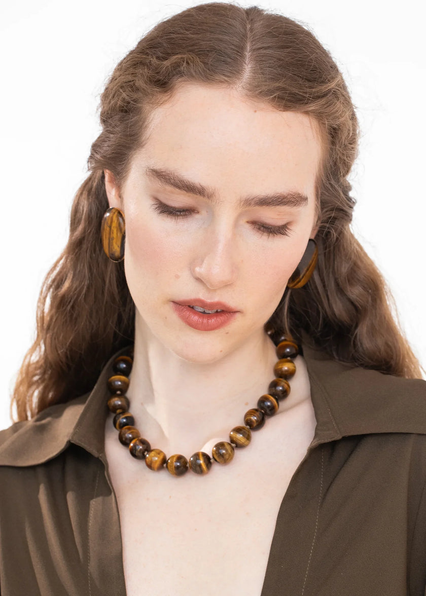 A woman with brown hair wearing a chunky brown and black tiger's eye beaded necklace and matching large round earrings, styled with an olive green shirt against a white background.

Jennifer Behr- Louisa Necklace - Tigers Eye. Available at Edit by Elaine Turner. Model View. 