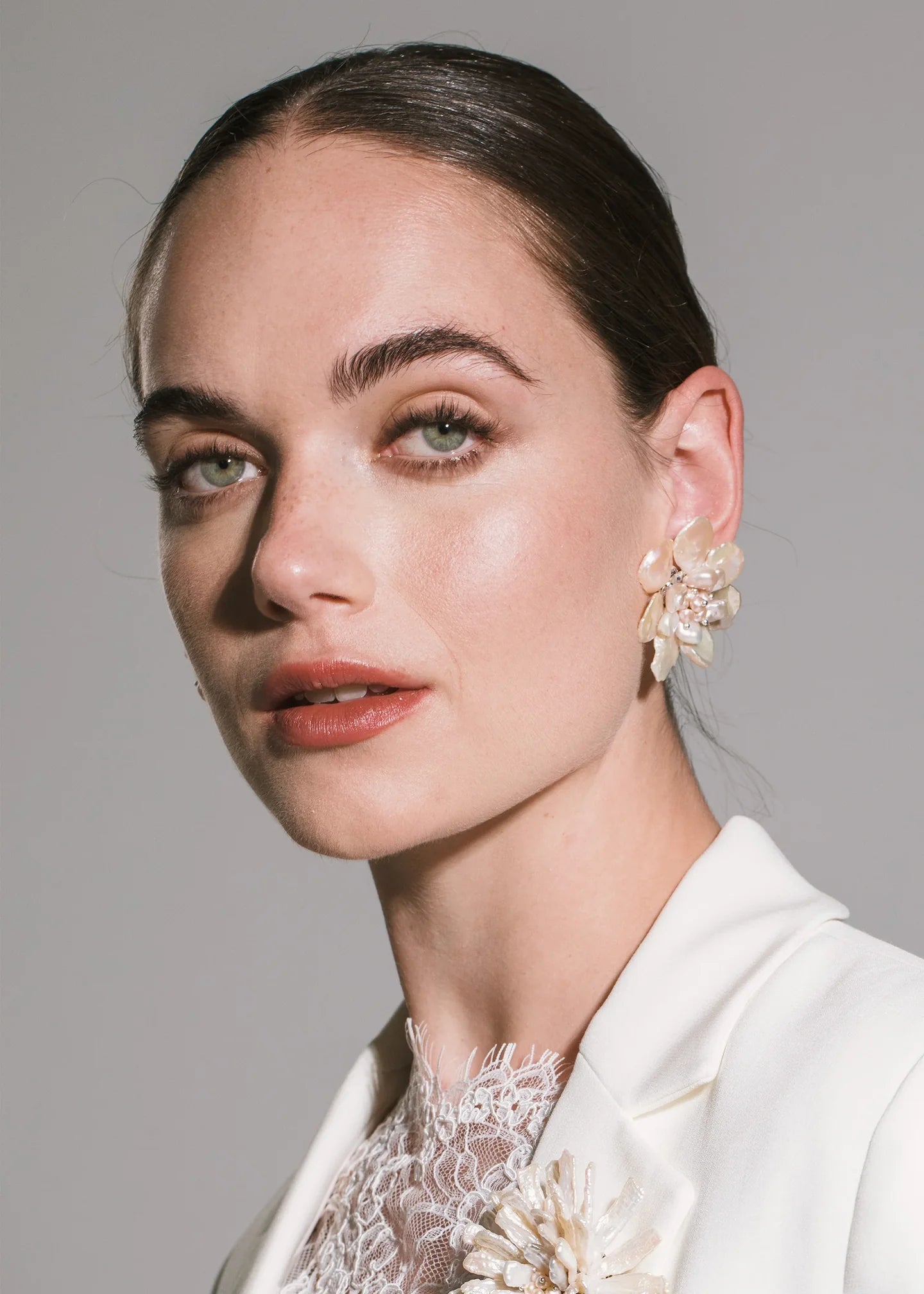 A close-up portrait of a woman with dark hair pulled back, wearing a white blazer and a white lace top, with a large floral gold statement earring visible on her left ear against a plain gray background.

Jennifer Behr- Margo Earrings - Pearl. Available at Edit by Elaine Turner in Houston, TX. Model View. 