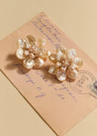 Two flower-shaped earrings made from iridescent pearl-like petals and small bead clusters, displayed on top of a vintage handwritten envelope against a warm beige background.

Jennifer Behr- Margo Earrings - Pearl. Available at Edit by Elaine Turner in Houston, TX. 
