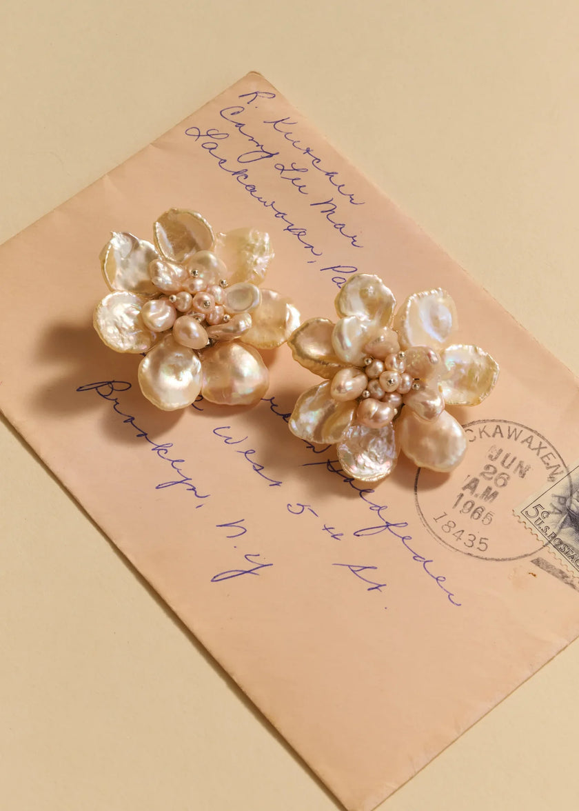 Two flower-shaped earrings made from iridescent pearl-like petals and small bead clusters, displayed on top of a vintage handwritten envelope against a warm beige background.

Jennifer Behr- Margo Earrings - Pearl. Available at Edit by Elaine Turner in Houston, TX. 