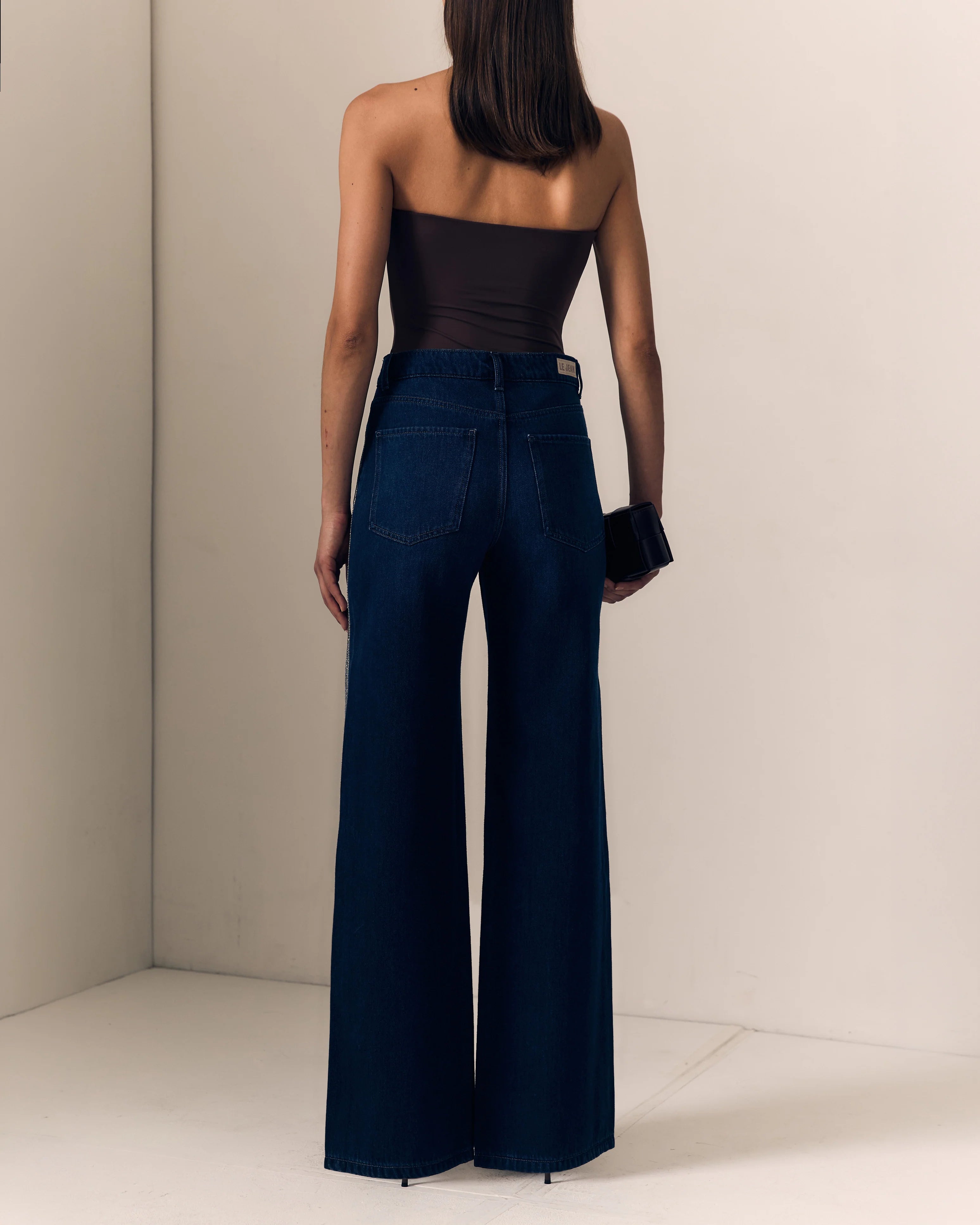 A woman shown from behind wearing dark blue wide-leg jeans and a black strapless top, standing in a minimalist beige studio corner.

Le Jean- Beaded Column in Midnight Rodeo. Available at Edit by Elaine Turner in Houston, TX. Model back view. 