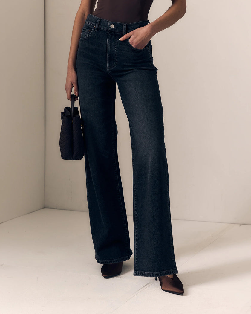 A model wearing dark blue wide-leg denim jeans, holding a small black handbag, standing in a minimalist beige room corner.

Le Jean- Column Jean - Fade to Black 
Model Close Up view. Available at Edit by Elaine Turner in Houston, TX. 