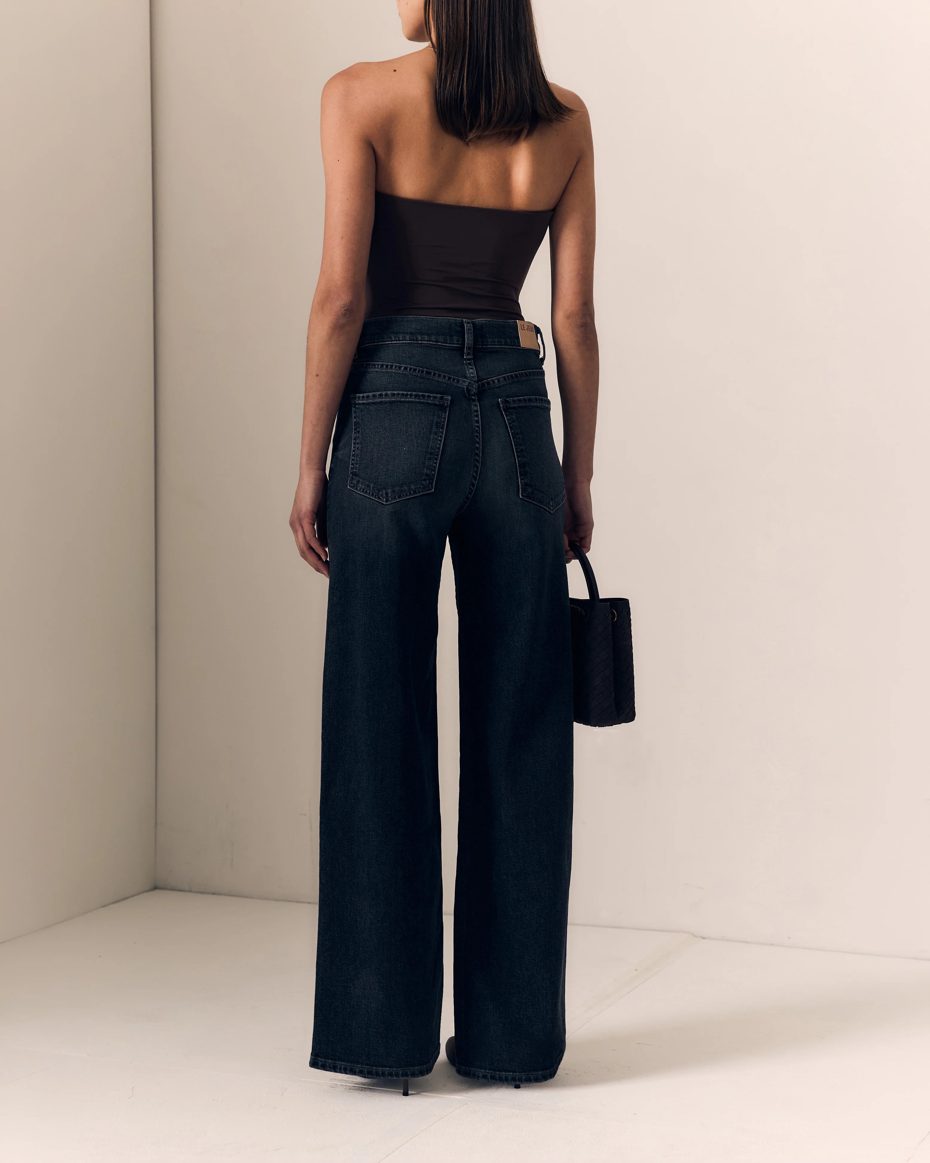 A model shown from behind wearing dark wide-leg denim jeans, a black strapless top, and holding a small black handbag in a minimalist beige room.

Le Jean- Column Jean - Fade to Black 
Full model back view. Available at Edit by Elaine Turner in Houston, TX. 