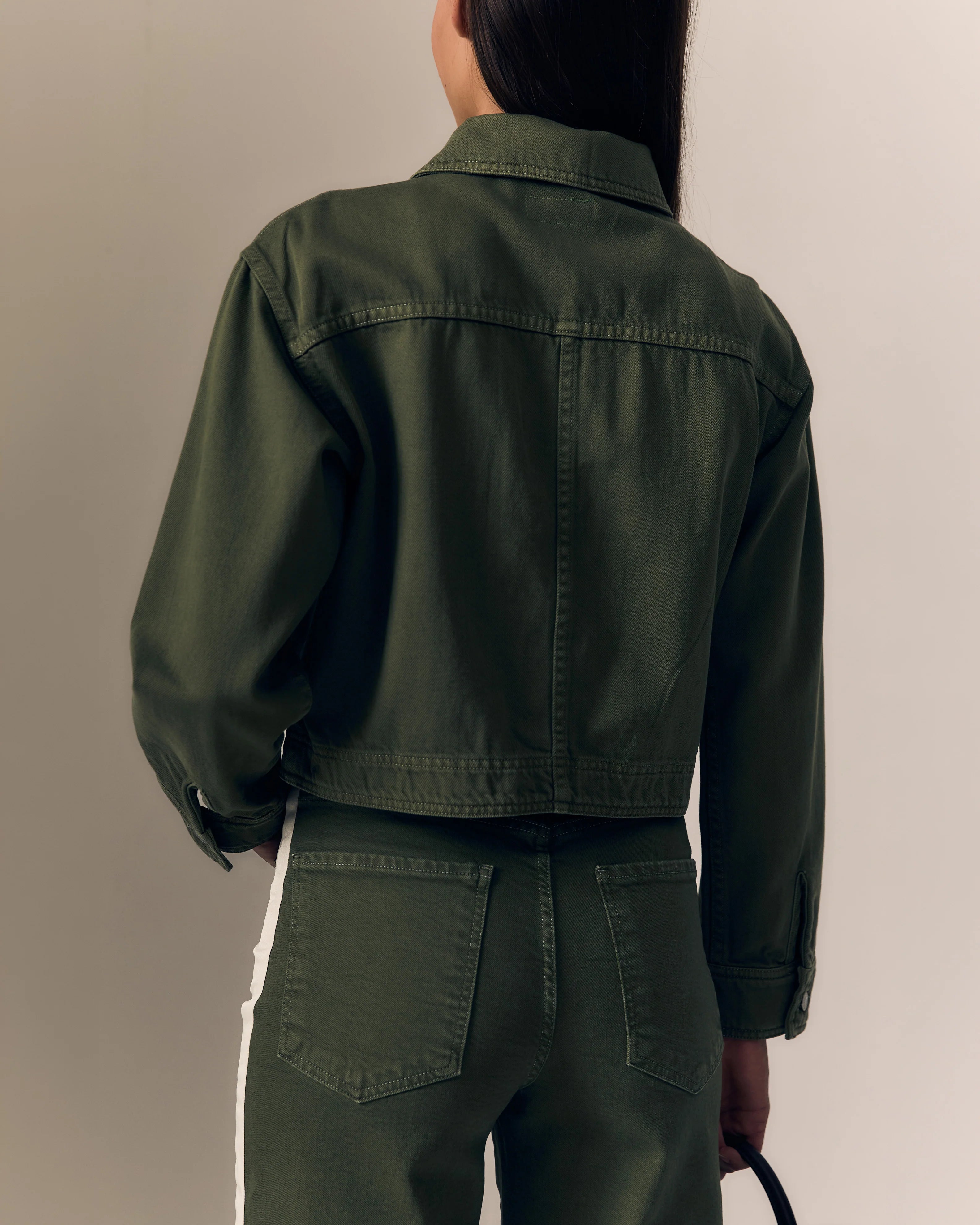 Person wearing a green jacket and pants against a neutral background. 

Le Jean Cora Jacket in olive green. Available at Edit by Elaine Turner in Houston, TX. Model Back Close-Up View.