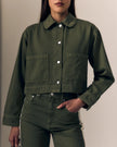 A woman wearing a cropped olive green utility jacket with a collar, silver snap buttons, and two large front patch pockets, paired with matching olive green jeans against a plain beige background.

Le Jean Cora Jacket in olive green. Available at Edit by Elaine Turner in Houston, TX. Model Front Close-Up View. 