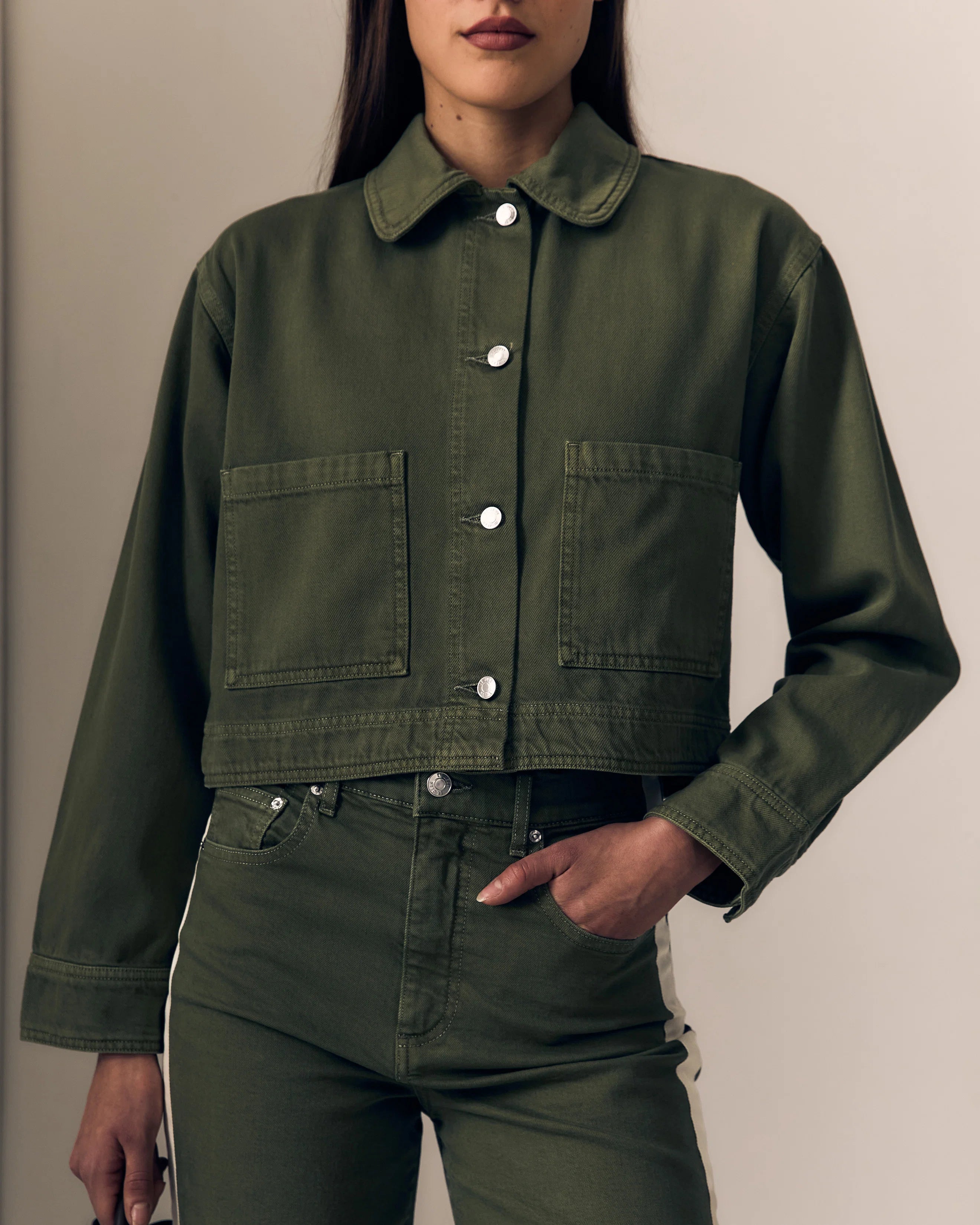 A woman wearing a cropped olive green utility jacket with a collar, silver snap buttons, and two large front patch pockets, paired with matching olive green jeans against a plain beige background.

Le Jean Cora Jacket in olive green. Available at Edit by Elaine Turner in Houston, TX. Model Front Close-Up View. 