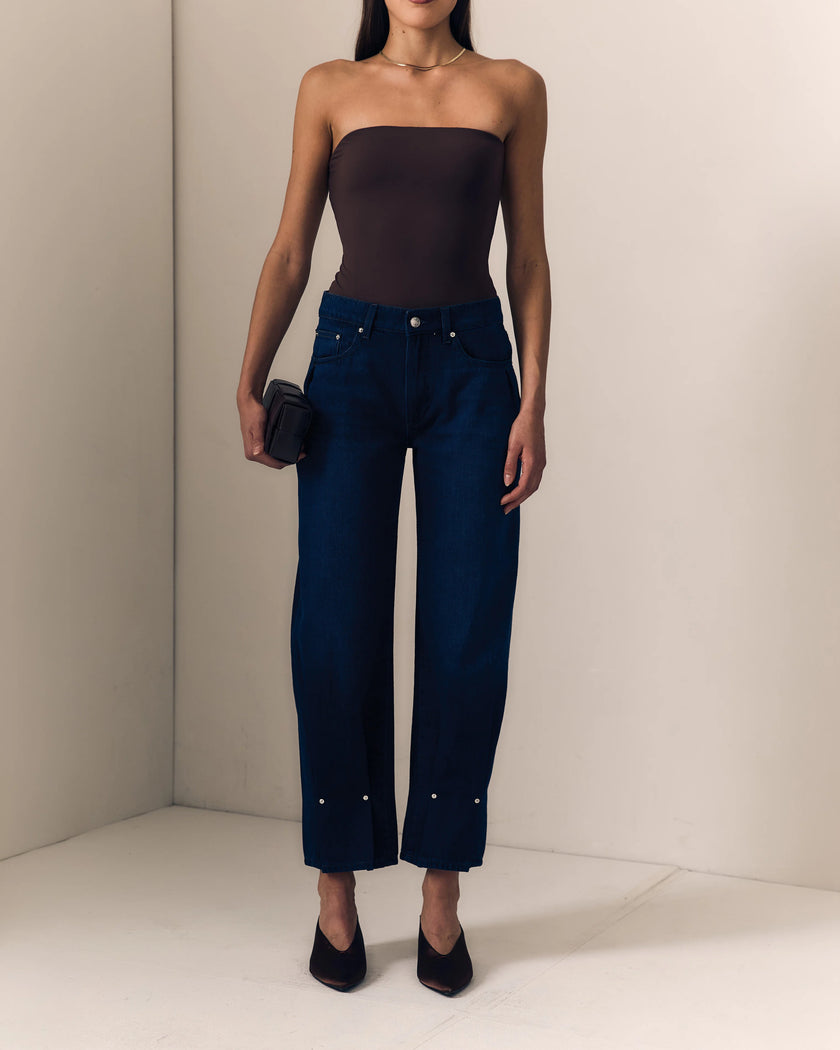 A model wearing a softly sculpted barrel leg jean with silver rivet details at the hem, paired with a dark brown strapless top and black heels, standing in a minimalist beige room.

Le Jean Delphine Jean in Midnight Rodeo. Available at Edit by Elaine Turner in Houston, TX. Full model front view. 