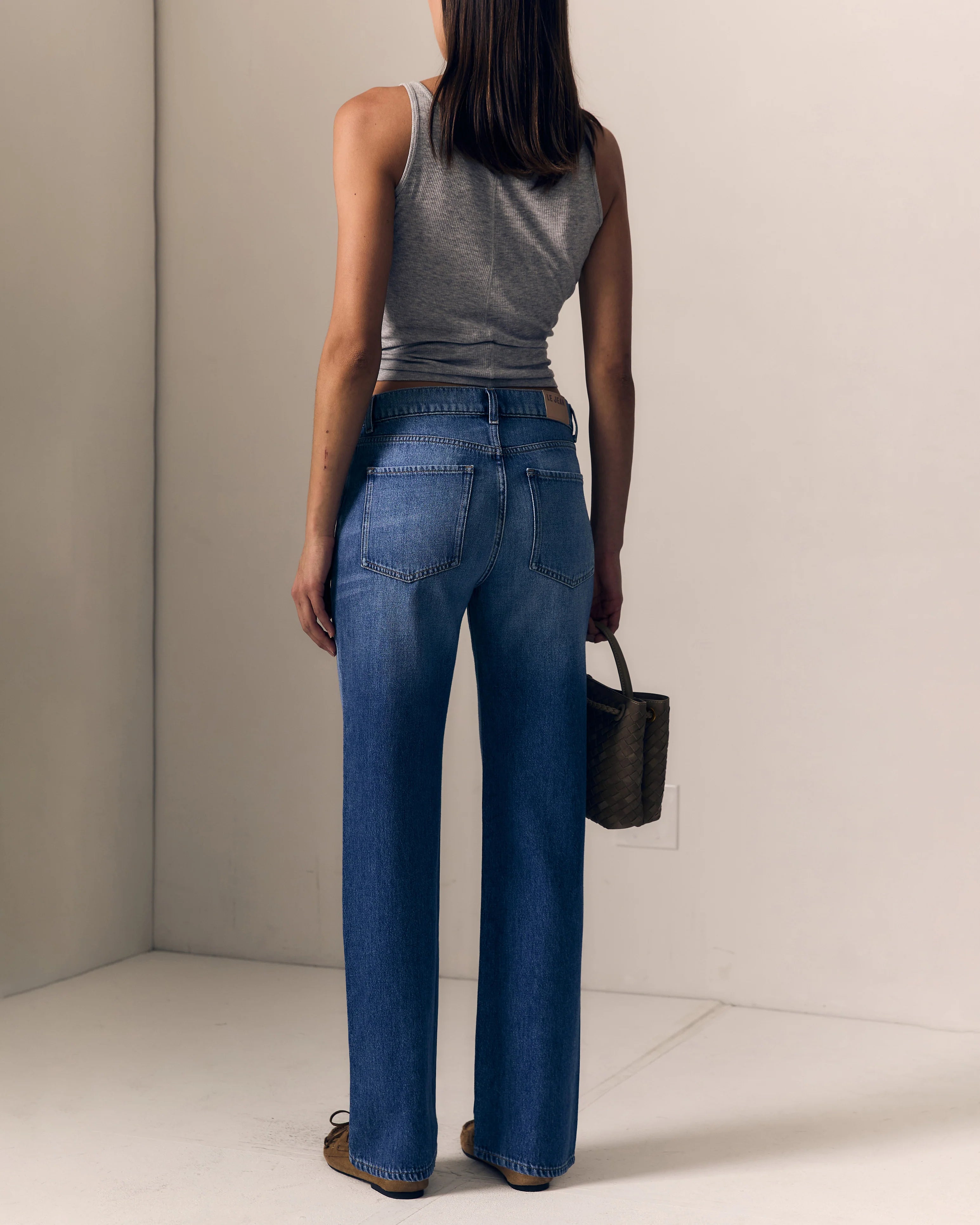 A woman is shown from behind wearing blue straight-leg jeans, a gray sleeveless top, and brown shoes, holding a small dark handbag in a minimalist studio setting.

Le Jean Mid 90's Straight in North Star. Available at Edit by Elaine Turner in Houston, TX. Full model back view. 